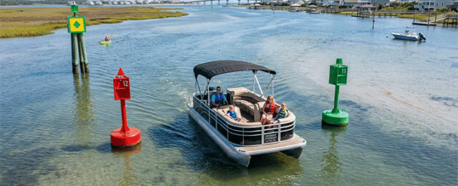 A family on a pontoon boat navigating a sunny, shallow channel on the Topsail Island Intracoastal Waterway, passing close to a red navigation buoy and green channel markers with the Surf City Bridge visible in the background.
